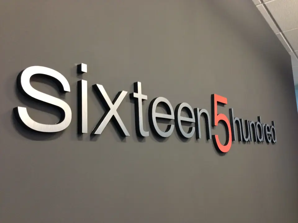 Brushed-metal dimensional letters spelling Sixteen5hundred on a gray office wall